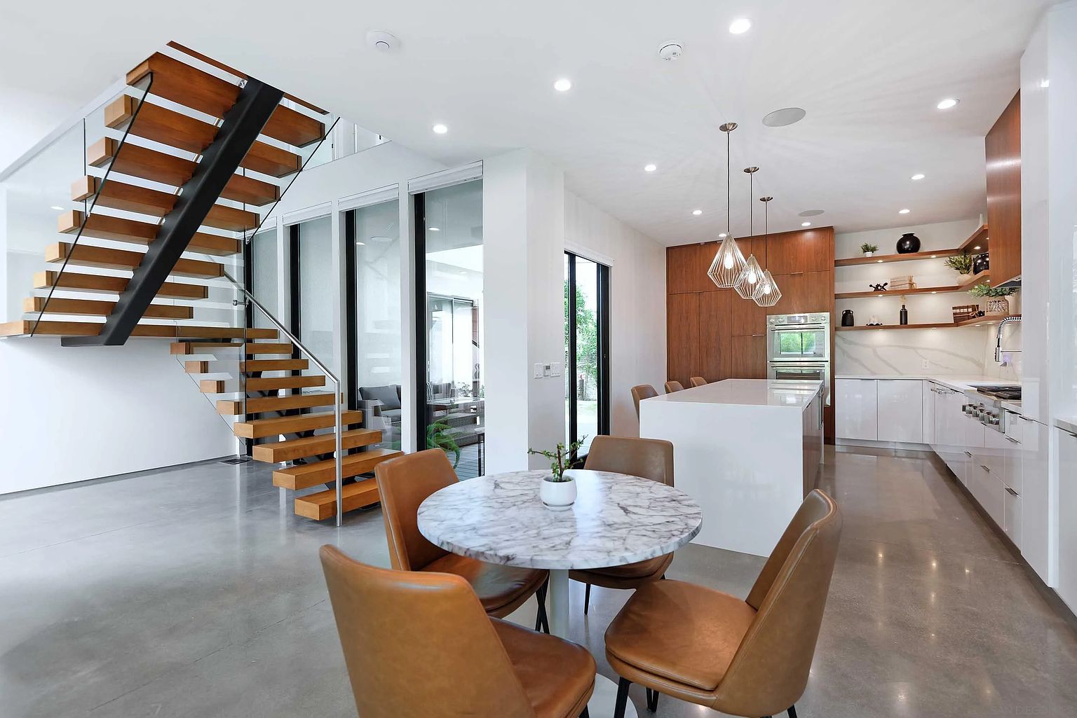 This interior shot showcases a modern dining area adjacent to a kitchen and staircase. A round marble-topped table is surrounded by four leather chairs, creating an intimate dining space. The open-concept design flows into a contemporary kitchen with sleek cabinetry and a central island, while a floating staircase adds architectural interest. The perspective is from a medium shot, highlighting the dining area and its connection to the surrounding spaces.