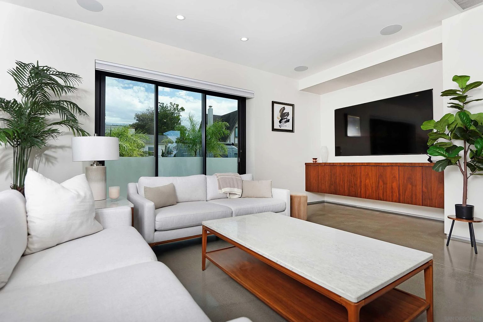 This is an interior shot of a modern living room. The room features a large window, a white sofa set, a marble-topped coffee table, and a wooden cabinet under a wall-mounted television. The style is minimalist and bright, creating a welcoming and spacious atmosphere.