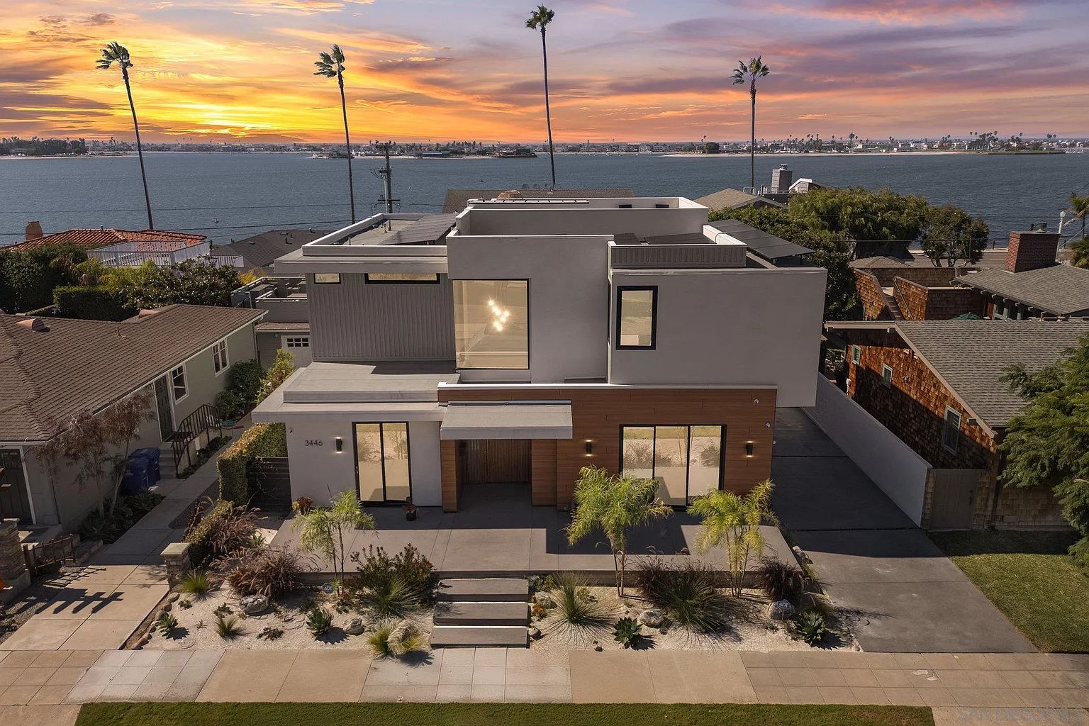 This is a front exterior view of a modern two-story home with a flat roof and a combination of white stucco and wood siding. The house features large windows and a minimalist landscape design with small trees and desert plants. The image captures a beautiful sunset over the water in the background, adding to the property's appeal.