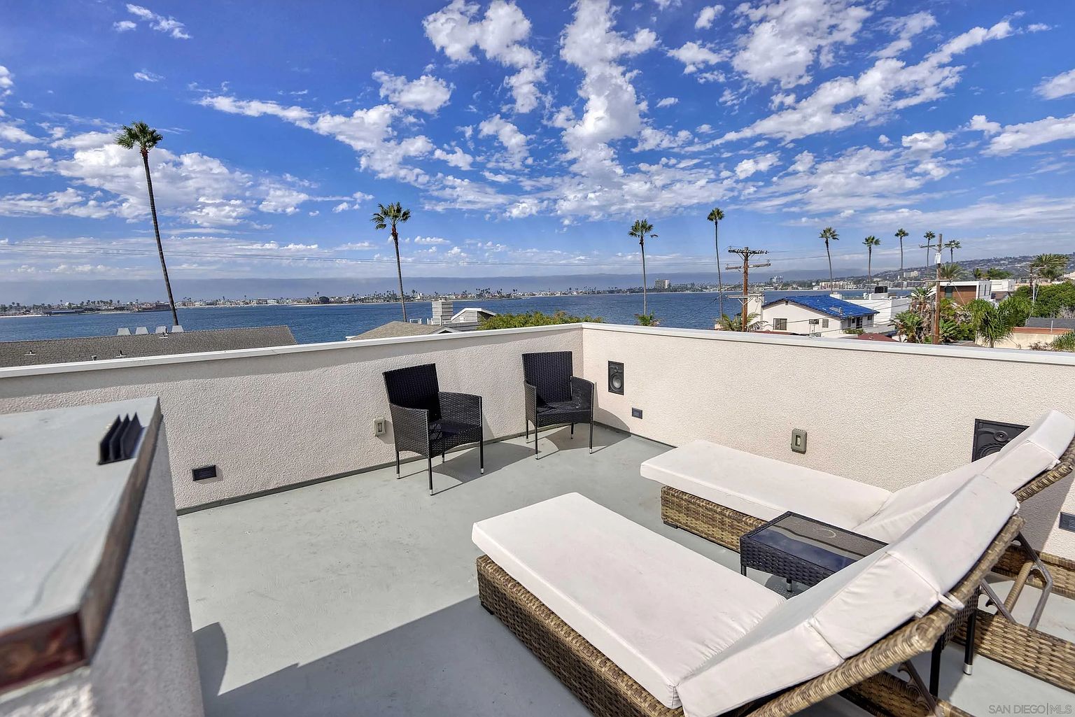 This image showcases a stylish rooftop patio or deck with a stunning view of the ocean and cityscape. The space is furnished with modern black chairs and comfortable white cushioned lounge chairs, creating an inviting atmosphere for relaxation and entertainment. The white stucco walls provide a clean backdrop, emphasizing the beautiful scenery and the upscale ambiance of the property.