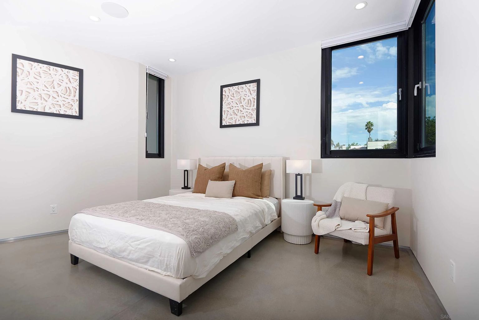 This is a bright and airy primary bedroom featuring a neutral color palette. The room includes a bed with decorative pillows and a throw, a side table with a lamp, and a comfortable chair. Natural light floods the space through a large window, enhancing the room's serene and inviting atmosphere.