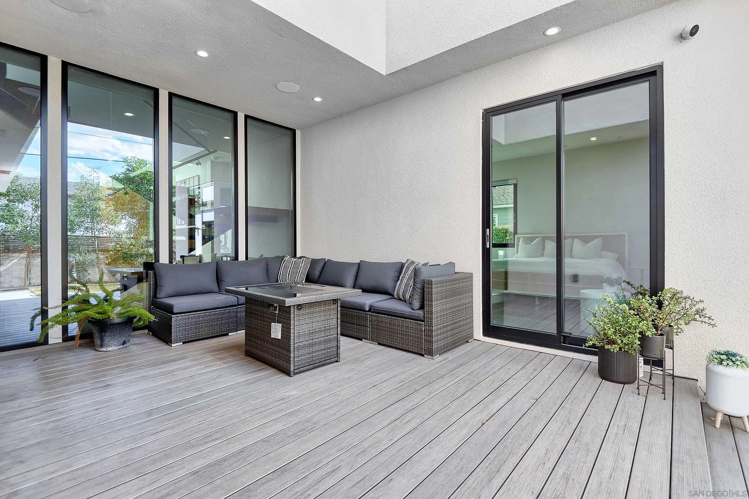 This image showcases a stylish outdoor patio area, featuring a modern gray wicker sectional sofa and a matching fire pit table. The deck is constructed with gray composite decking, and large windows and a sliding glass door provide access to the interior. The space is decorated with potted plants, creating a relaxing and inviting atmosphere.