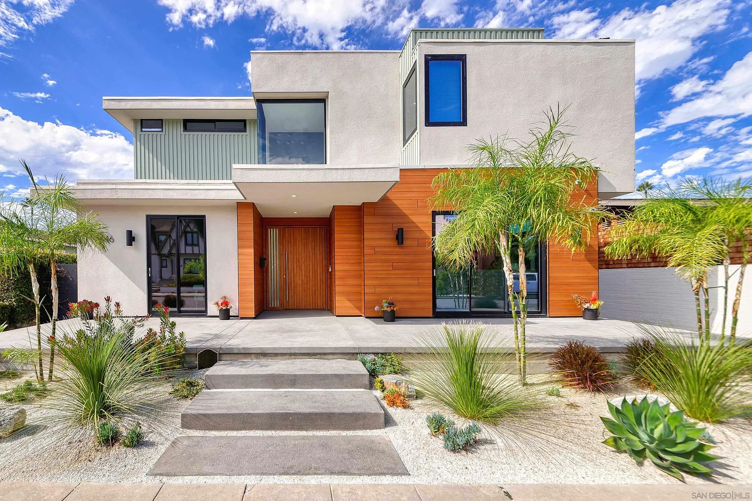 This is a front view of a modern two-story home with a striking architectural design. The exterior features a combination of light stucco, wood paneling, and black-framed windows, creating a contemporary aesthetic. The landscaping includes minimalist plantings with white gravel, adding to the clean and sophisticated curb appeal.
