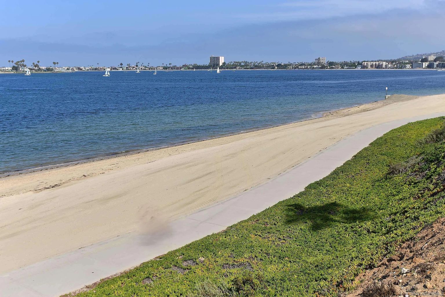 This image showcases a serene coastal scene with a sandy beach, calm blue water, and a paved pathway bordered by lush green vegetation. In the distance, buildings and sailboats dot the horizon, creating a picturesque backdrop. The view emphasizes the property's proximity to the beach and the potential for outdoor recreation and relaxation.