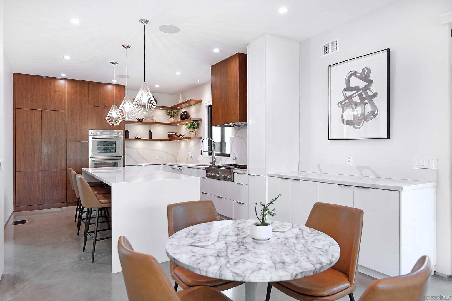 This is a modern kitchen with a clean and bright aesthetic. It features a marble-topped dining table with four leather chairs, a kitchen island with bar stools, and sleek white cabinetry complemented by wooden accents. The space is well-lit with recessed lighting and pendant lights, creating a welcoming and stylish atmosphere.