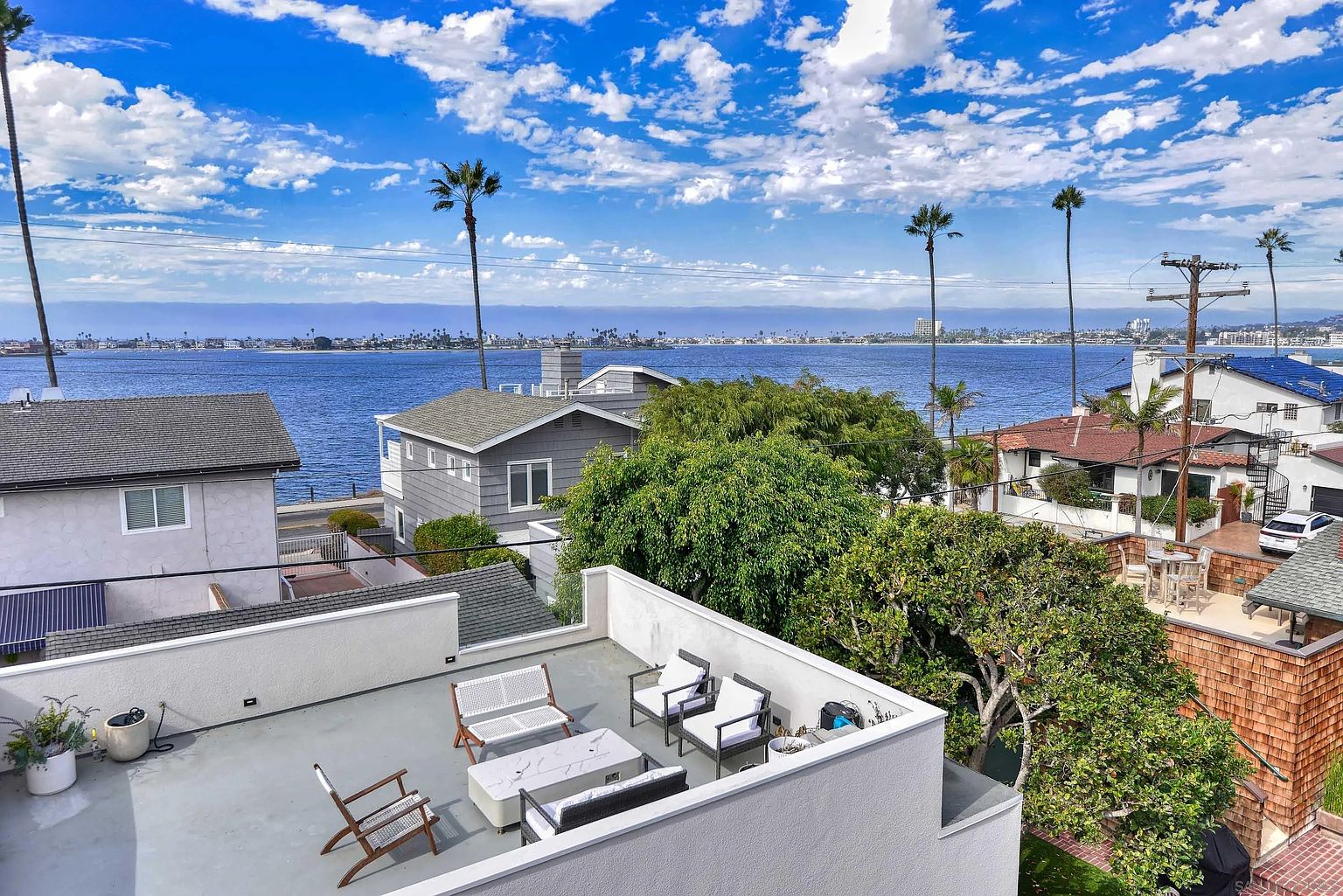 This image showcases a stylish rooftop patio with modern outdoor furniture, including a seating area with a fire pit. The patio offers stunning views of the ocean and cityscape, framed by palm trees and clear blue skies. The overall impression is one of luxury and relaxation, perfect for entertaining or enjoying the coastal scenery.