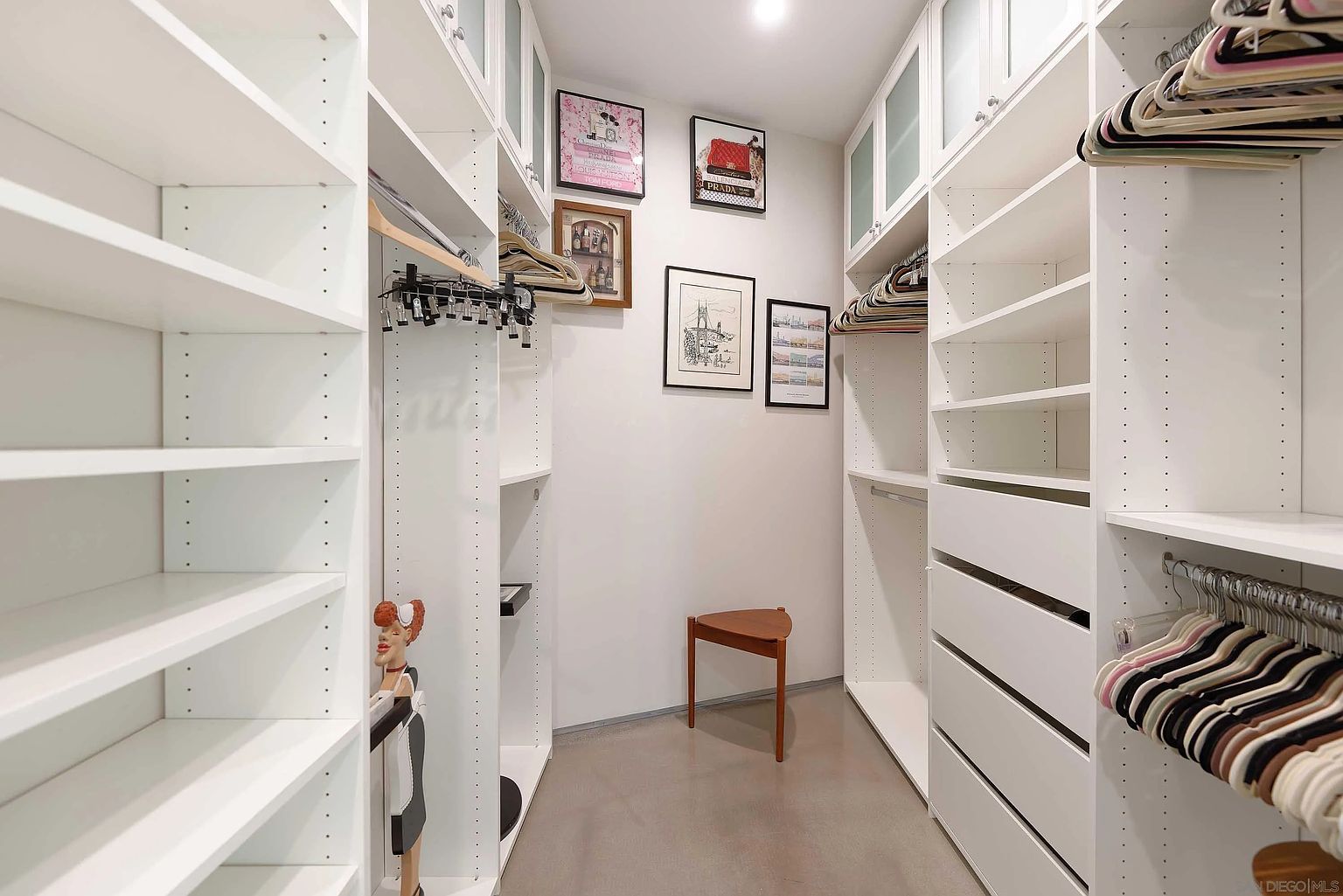This is a well-organized walk-in closet featuring custom white shelving and drawers on both sides of a narrow walkway. The closet is brightly lit, with decorative framed art pieces adorning the back wall, and a small triangular stool is placed in the center. The overall impression is clean, modern, and luxurious, highlighting ample storage space.