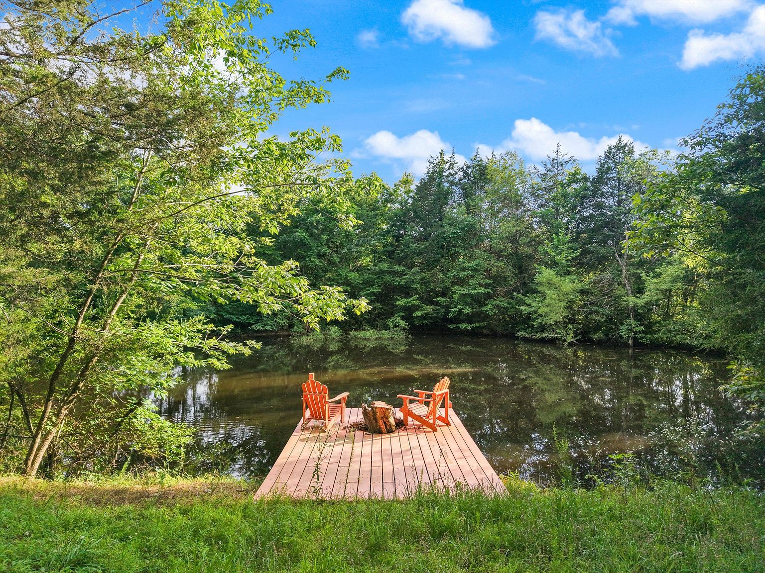This image showcases a serene backyard pond with a wooden dock featuring two Adirondack chairs and a small wooden table. The pond is surrounded by lush greenery and trees, creating a peaceful and private setting. The scene evokes a sense of relaxation and natural beauty, highlighting the property's outdoor amenities.