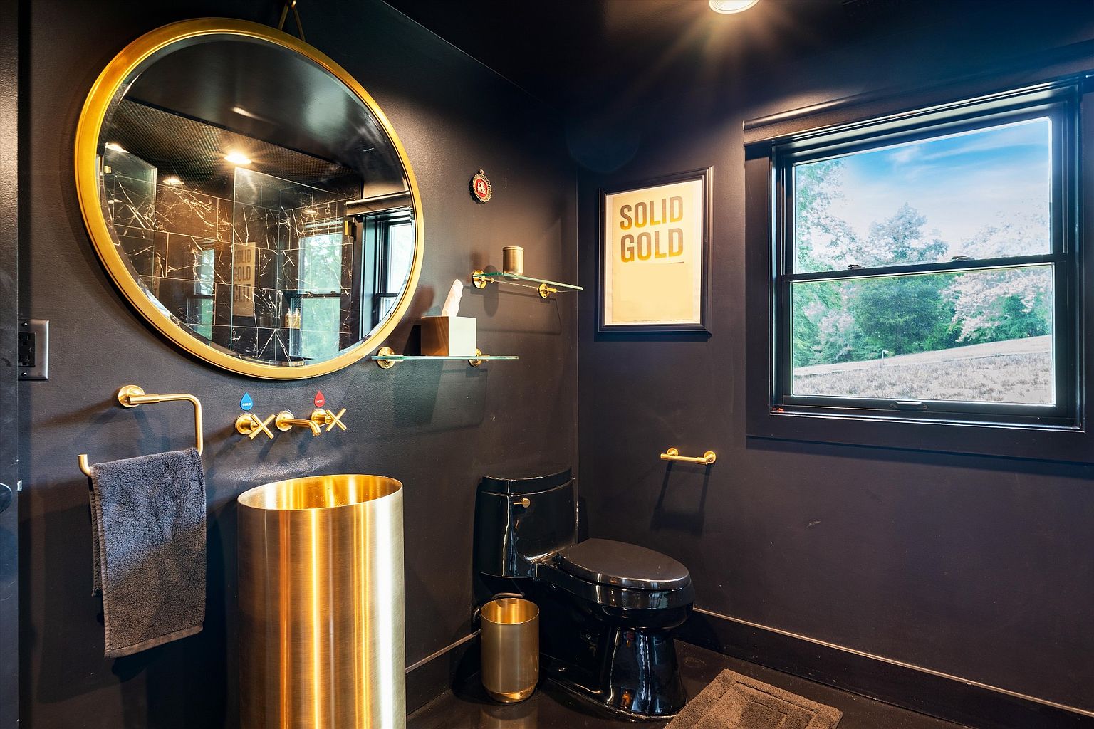 This is a modern bathroom with a bold, dark aesthetic. The walls are painted a deep black, contrasted by gold fixtures, including a round mirror, faucet, and cylindrical sink. A black toilet and a framed "SOLID GOLD" print add to the luxurious and stylish ambiance.