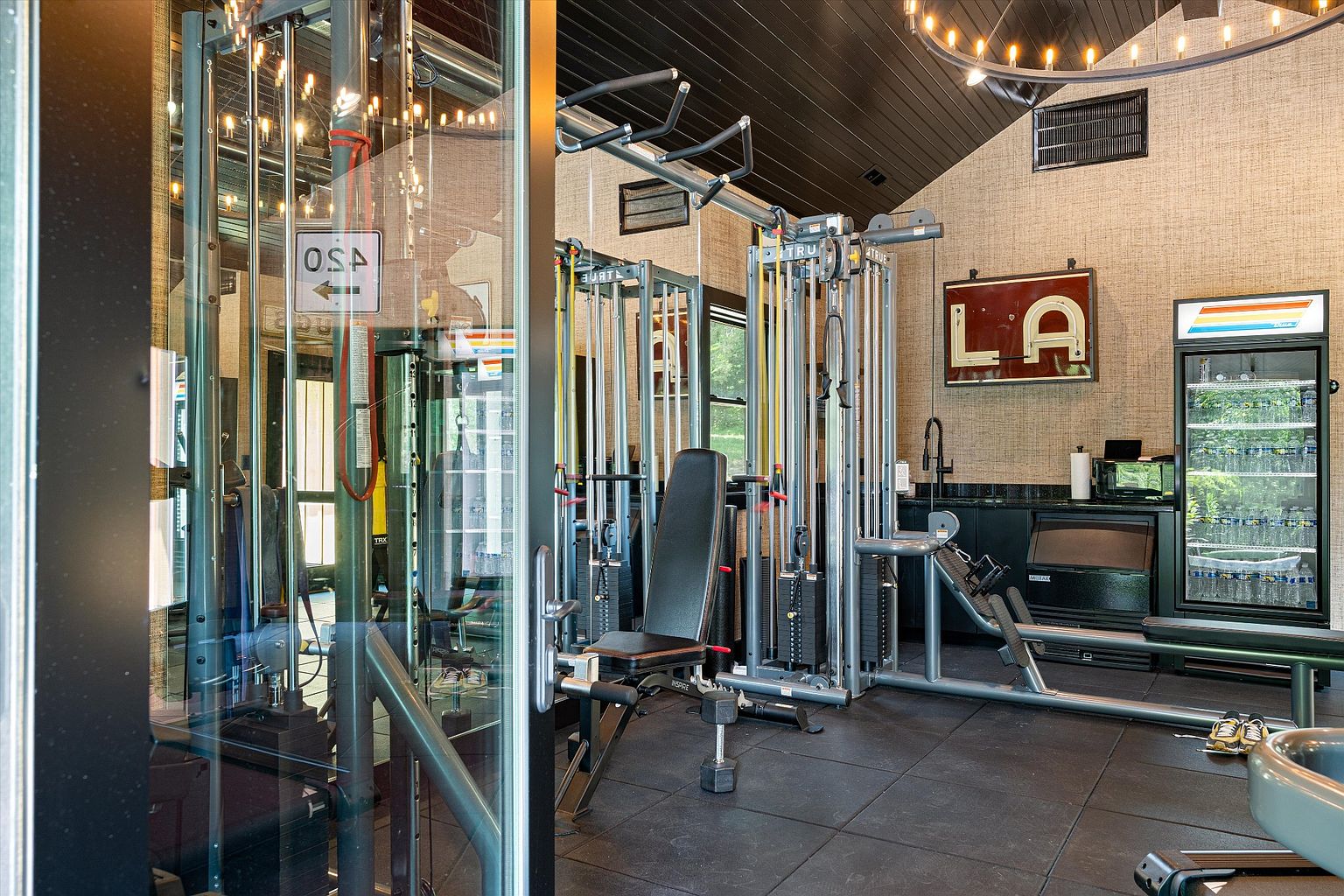 This is a well-equipped home gym featuring various weight machines, including a cable machine and a leg press. The room has a dark tile floor, textured wallpaper, and a black ceiling with a modern light fixture. A beverage refrigerator and a small sink area are also present, adding convenience to the workout space.