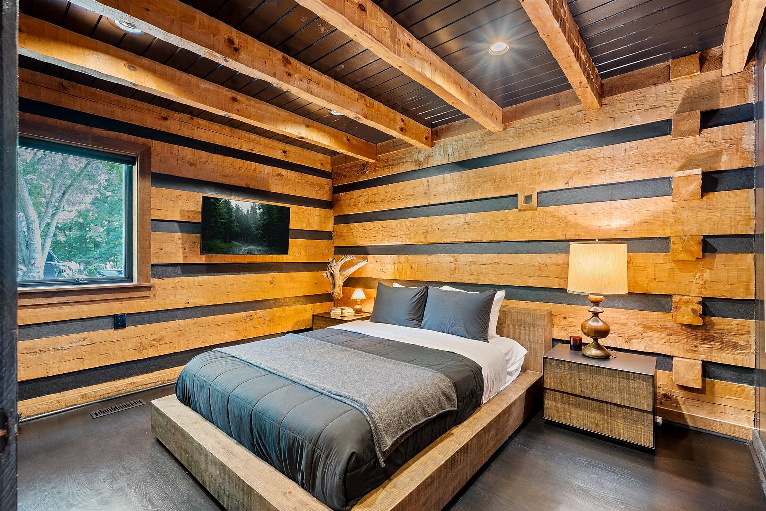 This is a cozy primary bedroom featuring a unique log cabin style with exposed wooden beams and dark accents. The room is furnished with a platform bed, a nightstand with a lamp, and a wall-mounted television. The dark wood flooring and ceiling add to the rustic yet modern aesthetic.