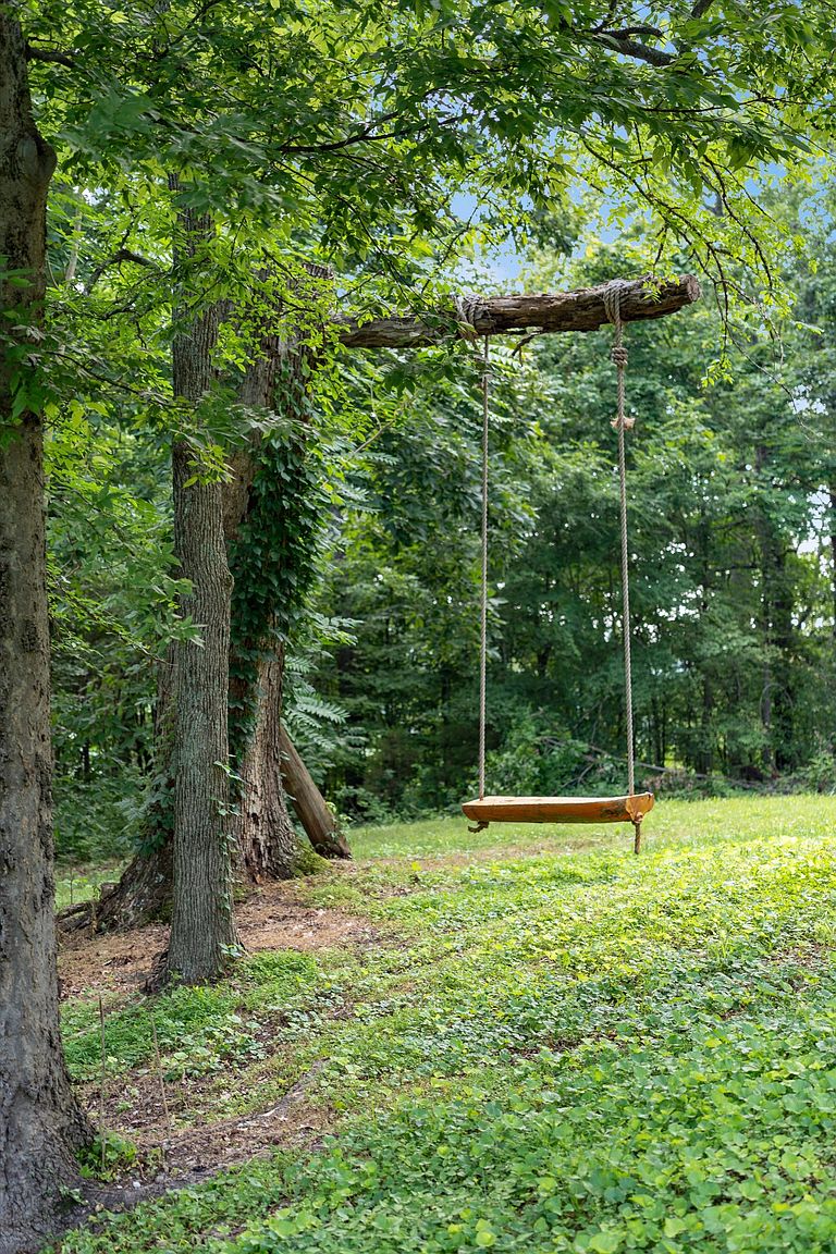 This image showcases a charming yard or garden featuring a rustic wooden swing suspended from a tree branch. The swing is the focal point, surrounded by lush greenery and mature trees, creating a serene and inviting atmosphere. The scene evokes a sense of tranquility and natural beauty, perfect for relaxation and outdoor enjoyment.