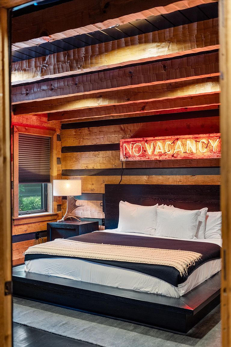 This is a cozy primary bedroom featuring a rustic, log cabin style with exposed wooden beams and a "NO VACANCY" neon sign. The bed is a modern platform design with a dark wood frame, crisp white linens, and a dark blanket with a textured throw. A window with blinds provides natural light, and a unique antler-based lamp sits on the bedside table.