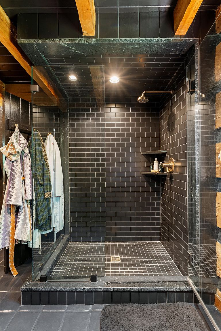 This is a modern primary bathroom featuring a walk-in shower with black subway tile walls and a grid-patterned floor. The shower has glass doors, a rainfall showerhead, and a built-in shelf for toiletries. The overall impression is sleek and luxurious, with a focus on dark, sophisticated tones.