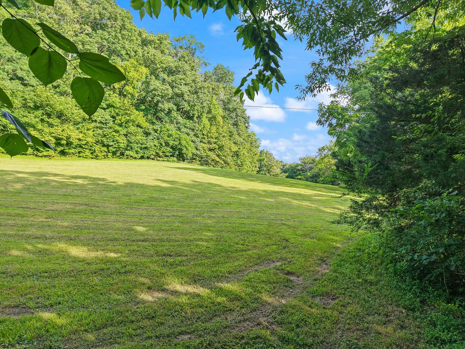 The image showcases a large, well-maintained grassy yard or field, bordered by lush green trees and foliage. The landscape slopes gently upwards, creating a sense of spaciousness and tranquility. The scene is bathed in natural light, suggesting a peaceful and inviting outdoor space, ideal for recreation or relaxation.