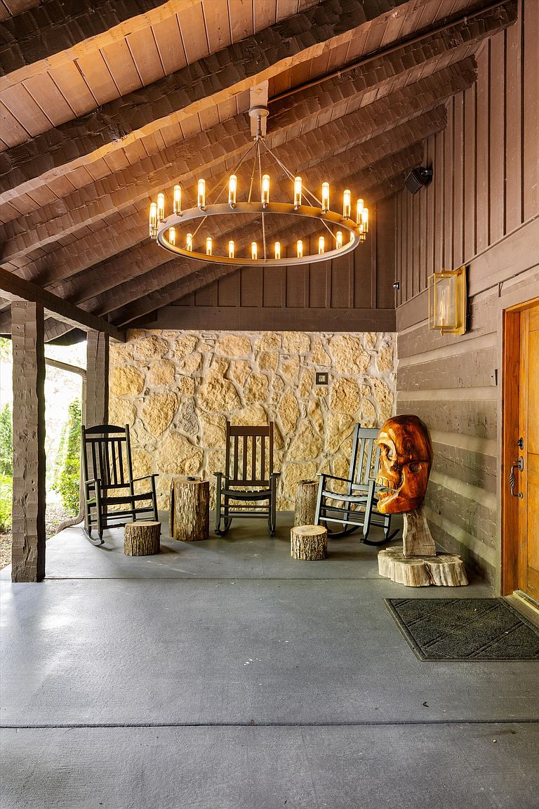This image showcases a rustic and inviting covered patio area. The space features a stone accent wall, wooden rocking chairs, and unique log stump side tables, all illuminated by a striking circular chandelier. A wooden skull sculpture adds an artistic touch, enhancing the area's character and charm.