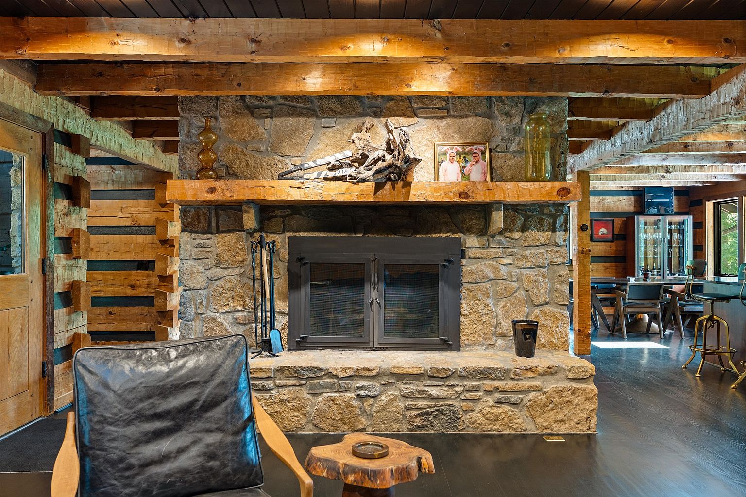 This is an interior shot of a rustic living room featuring a stone fireplace as the focal point. The room has exposed wooden beams on the ceiling and log-style walls, contributing to a warm and inviting atmosphere. A leather chair sits in the foreground, suggesting a comfortable space for relaxation.