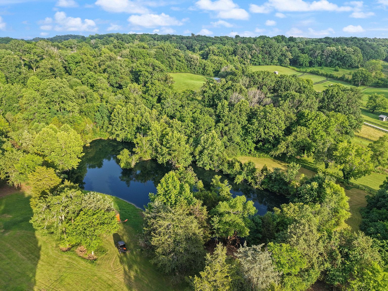 This aerial shot showcases a lush property featuring a serene pond surrounded by dense, mature trees. A well-maintained lawn extends to the water's edge, and a vehicle is parked nearby, suggesting easy access. The overall impression is one of privacy and natural beauty, ideal for a secluded estate.