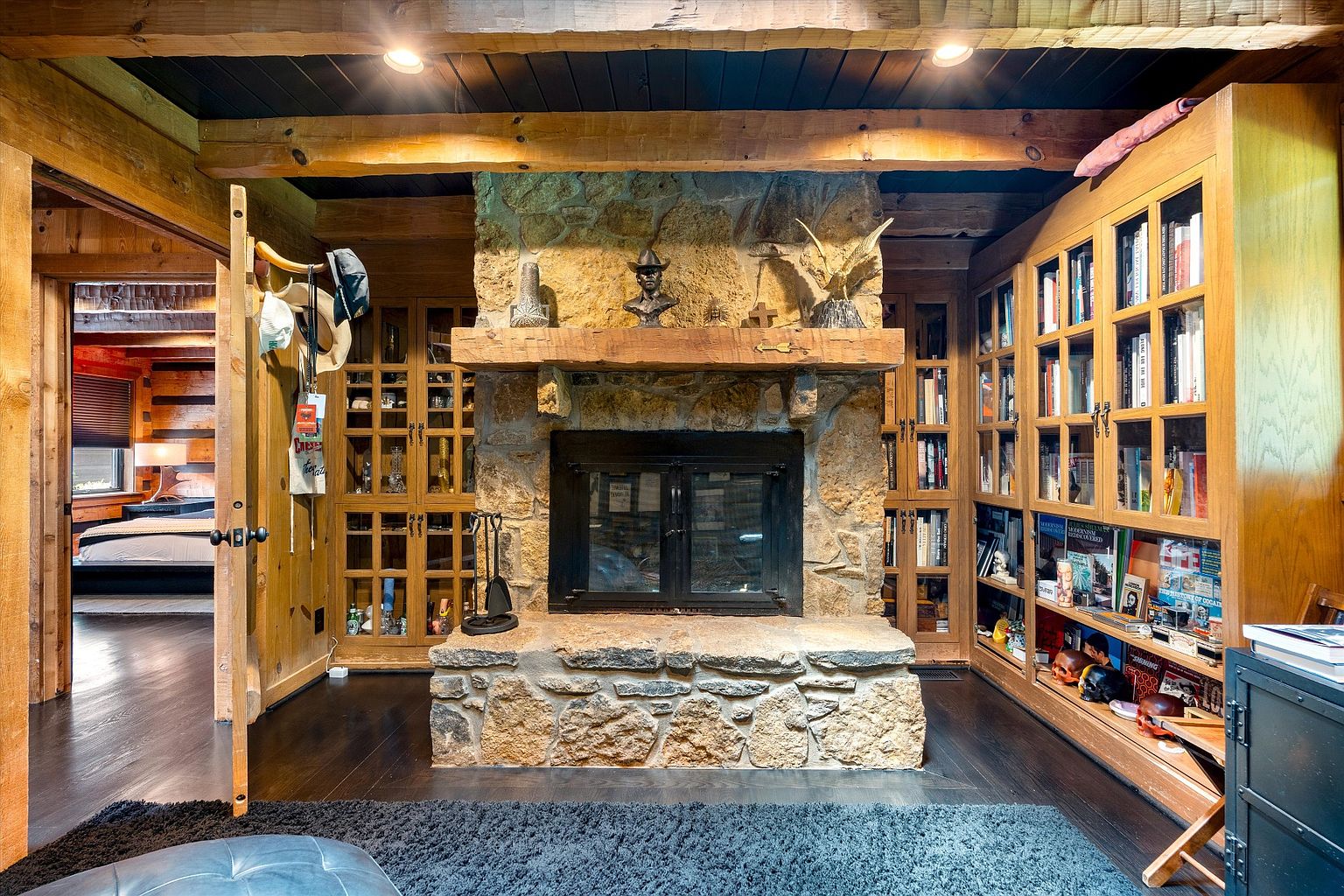 This interior shot showcases a cozy living room with a prominent stone fireplace as its focal point, flanked by built-in wooden bookshelves filled with books and decorative items. The room features dark hardwood floors, exposed wooden beams on the ceiling, and an open doorway leading to another room, creating a warm and inviting atmosphere. The perspective is from a low angle, emphasizing the height of the fireplace and the depth of the room.