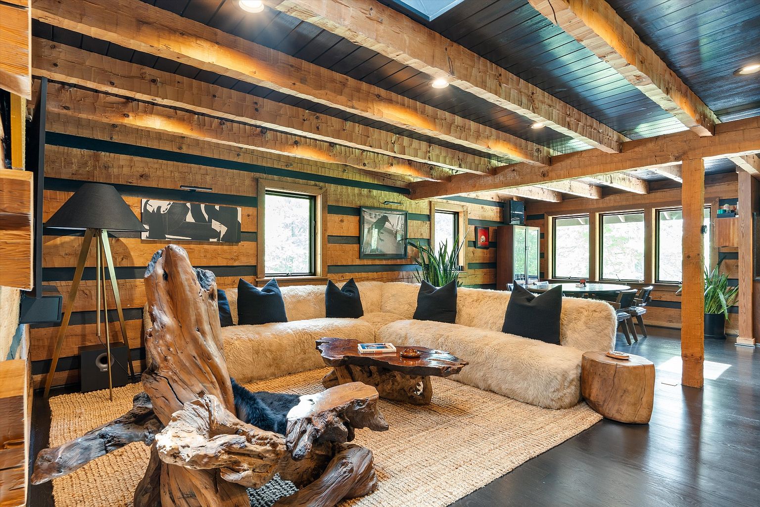 This is an interior shot of a rustic living room featuring exposed wooden beams and log walls, creating a warm and inviting atmosphere. A large, comfortable sectional sofa with dark pillows sits on a jute rug, complemented by unique wooden furniture pieces, including a coffee table and a sculptural chair made from tree trunks. Natural light streams in through multiple windows, enhancing the room's cozy and natural aesthetic.