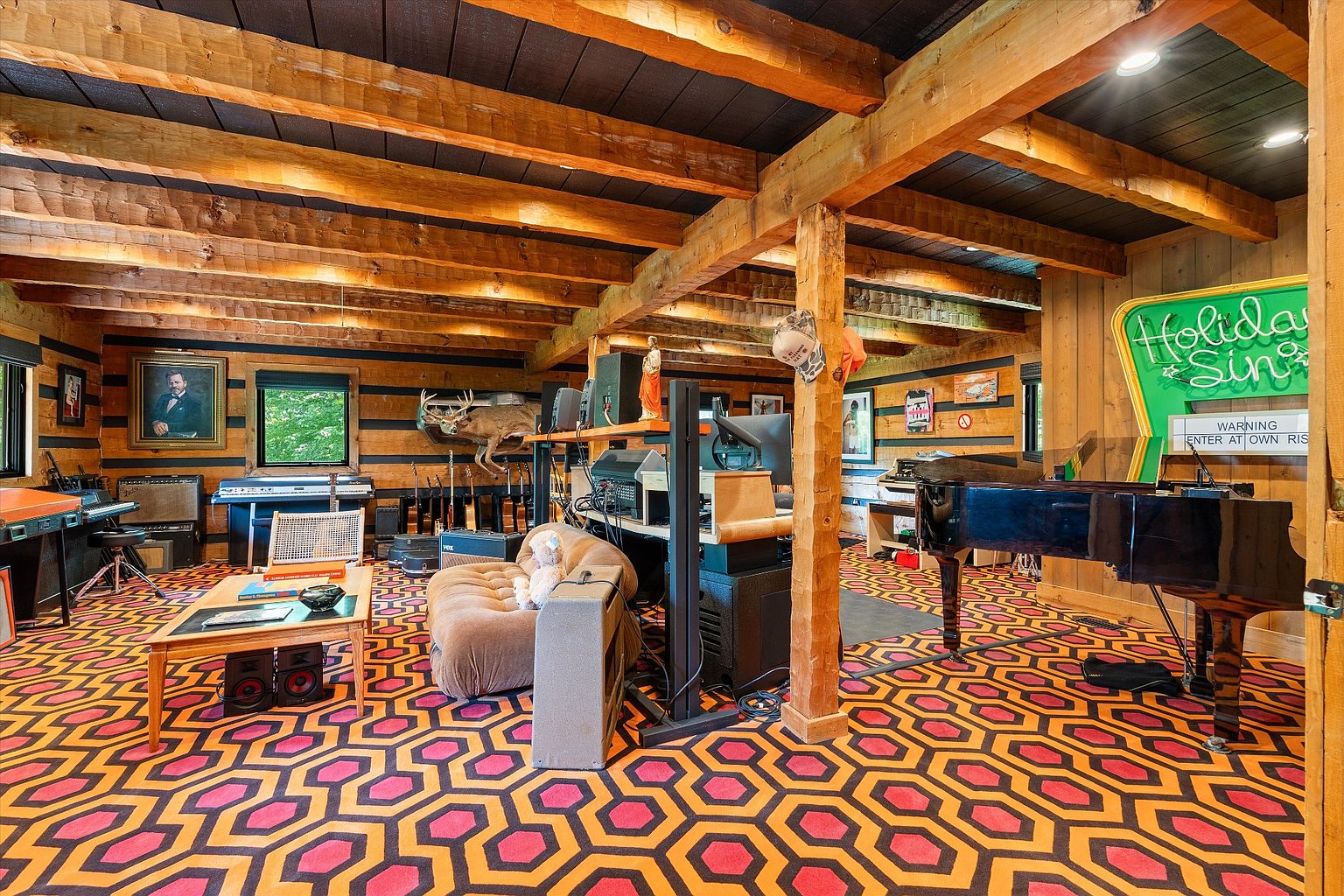 This unique living room features a striking geometric patterned carpet in orange and red, complemented by exposed wooden beams and a dark ceiling. The room is filled with musical instruments, including a grand piano and various keyboards, creating a creative and eclectic atmosphere. A comfortable armchair and a small coffee table add to the room's functionality and charm.