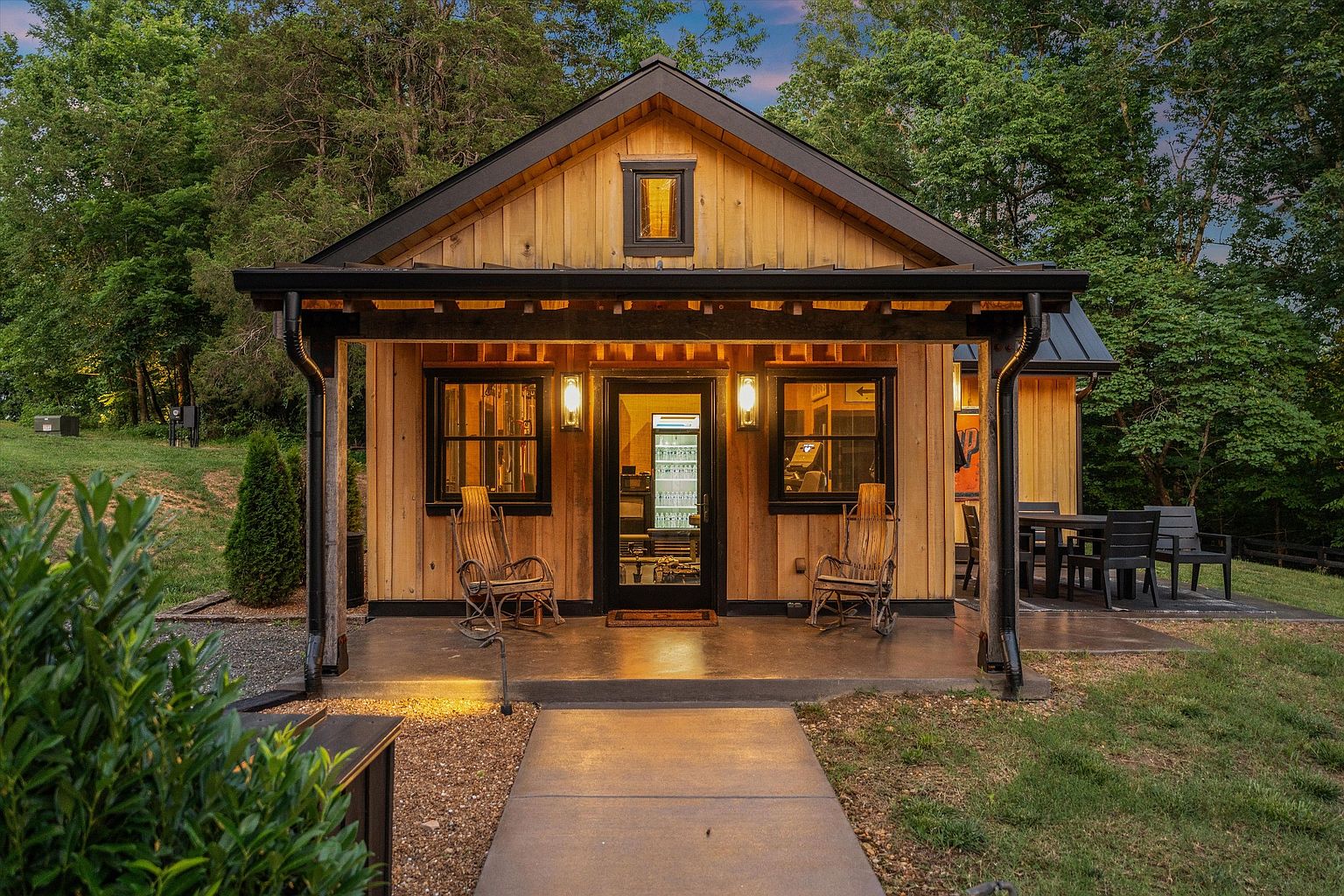 The image showcases the front exterior of a charming wooden cabin-style building. It features a covered porch with rocking chairs, a black-framed glass door, and symmetrical windows with black frames. The building is surrounded by lush greenery, creating a serene and inviting atmosphere, perfect for a rustic retreat.