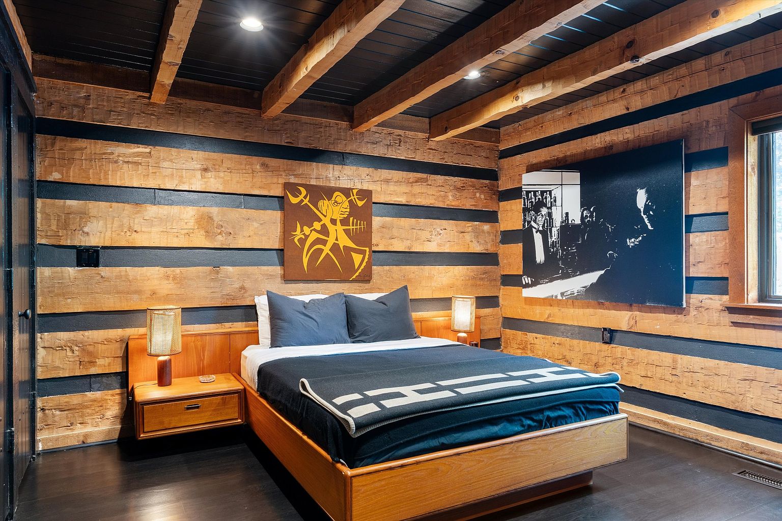 This is a primary bedroom featuring a unique log cabin style with dark-stained wooden beams and a black ceiling. The room is furnished with a wooden platform bed, matching nightstands, and artwork on the walls. The overall aesthetic is rustic yet modern, creating a cozy and stylish retreat.