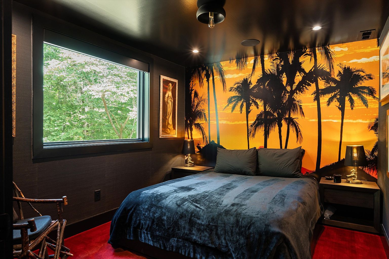 This is a bedroom featuring a dark, moody aesthetic with a large bed covered in a dark blue comforter and gray pillows. The wall behind the bed is adorned with a vibrant sunset mural featuring palm trees, creating a tropical contrast to the room's darker tones. A window to the left offers a view of lush greenery, and the room is furnished with bedside tables and lamps, adding to the ambiance.