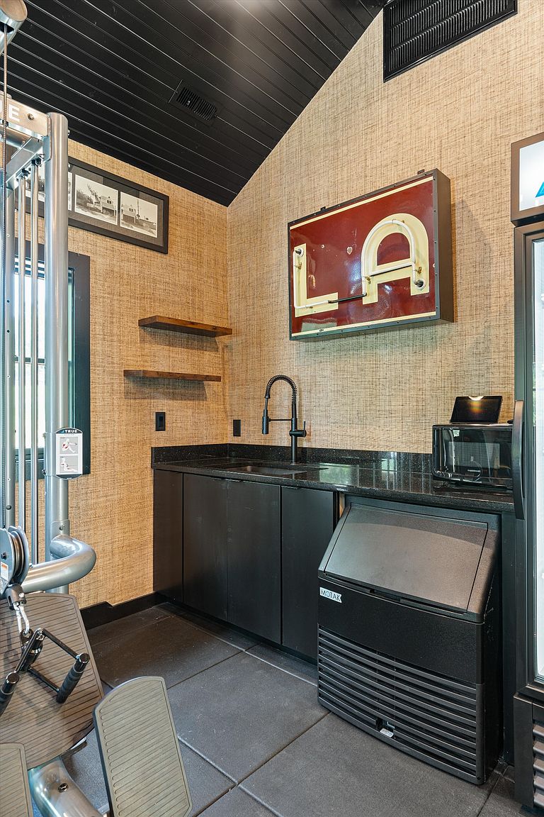 This image showcases a well-equipped home gym area featuring a multi-station weight machine, a wet bar with a black countertop and cabinets, and an ice maker. The walls are covered in textured wallpaper, and a vintage "LA" sign adds a unique decorative touch. The flooring appears to be rubberized tiles, providing a safe and functional workout space.