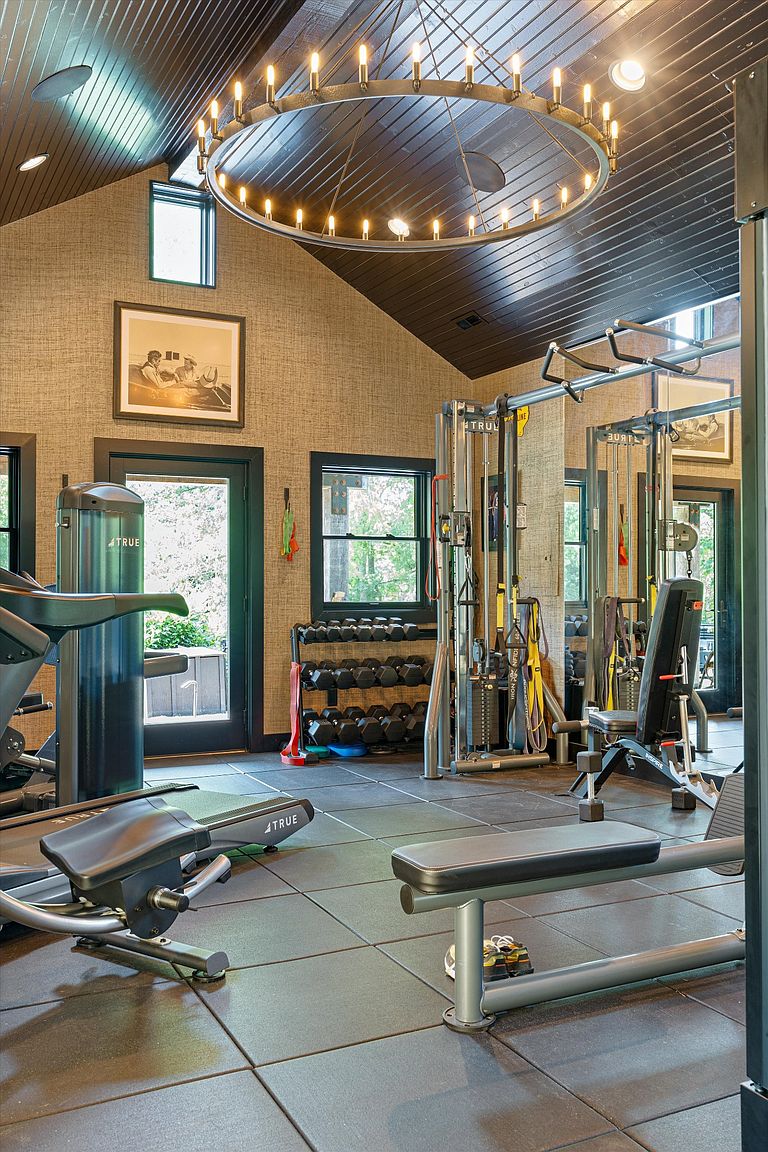 This is an interior shot of a well-equipped home gym. The room features a variety of exercise machines, including a treadmill and weightlifting stations, set on a black rubber tile floor. A large, circular chandelier hangs from the dark wood-paneled ceiling, providing ample lighting, and a framed picture adds a touch of sophistication to the textured walls.