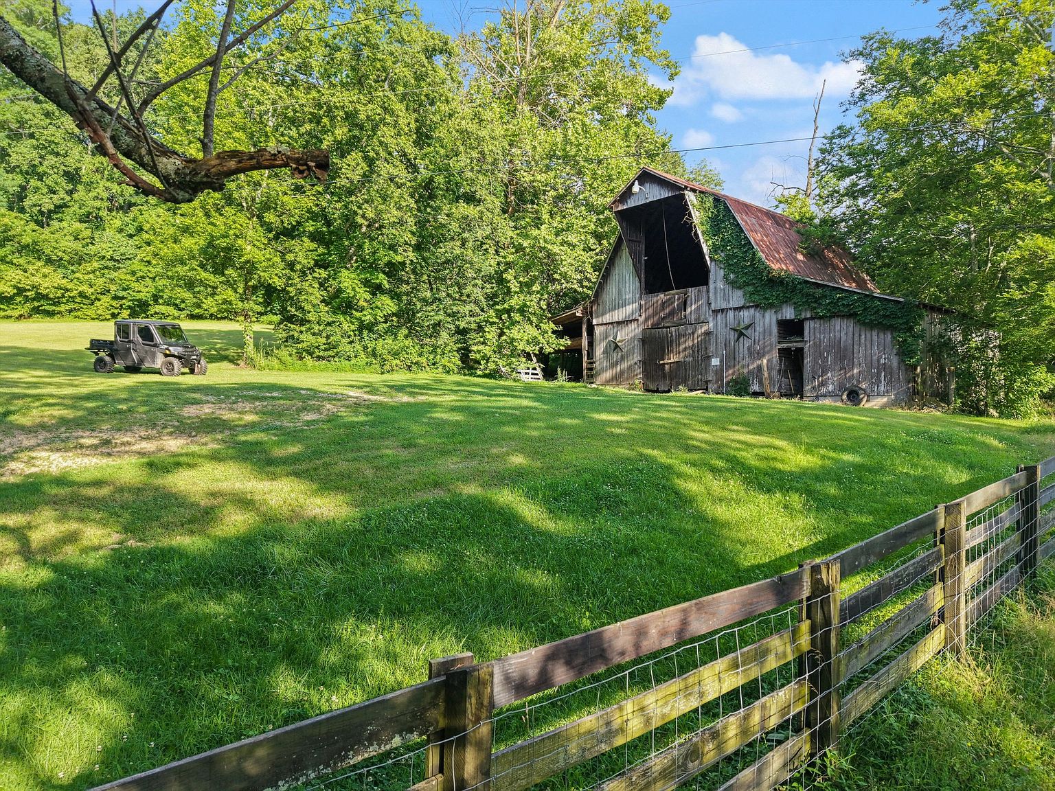 This exterior shot showcases a rustic barn with a weathered facade and a partially overgrown roof, set against a backdrop of lush greenery and a clear blue sky. A well-maintained lawn stretches out in front, bordered by a wooden fence, and a utility vehicle is parked in the distance, adding a touch of rural charm to the property. The scene evokes a sense of tranquility and timelessness.
