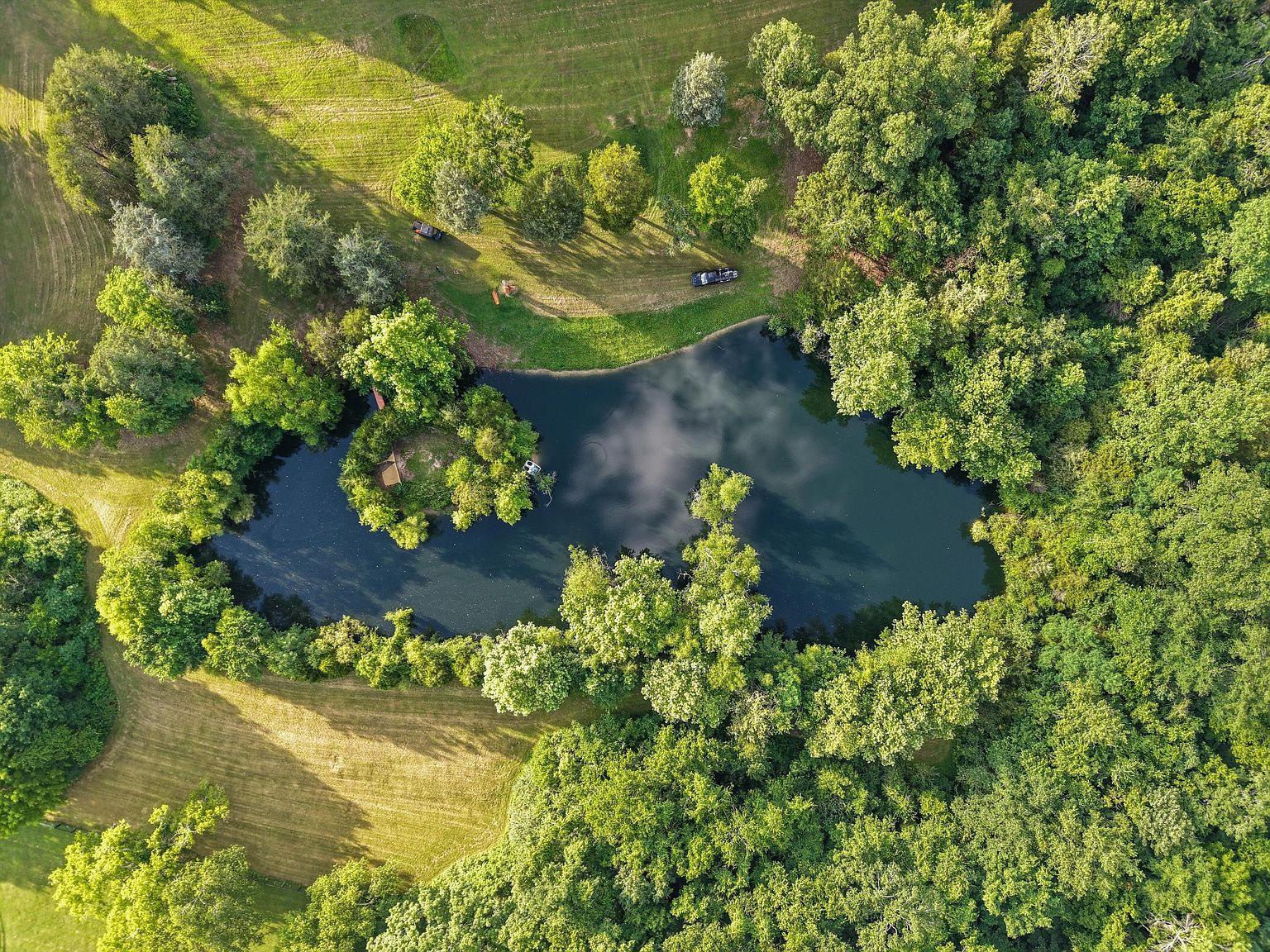 This aerial view showcases a serene property featuring a pond surrounded by lush greenery and mature trees. A small island with a structure sits in the middle of the pond, adding a unique focal point. The landscape includes manicured lawns and wooded areas, creating a private and picturesque setting.