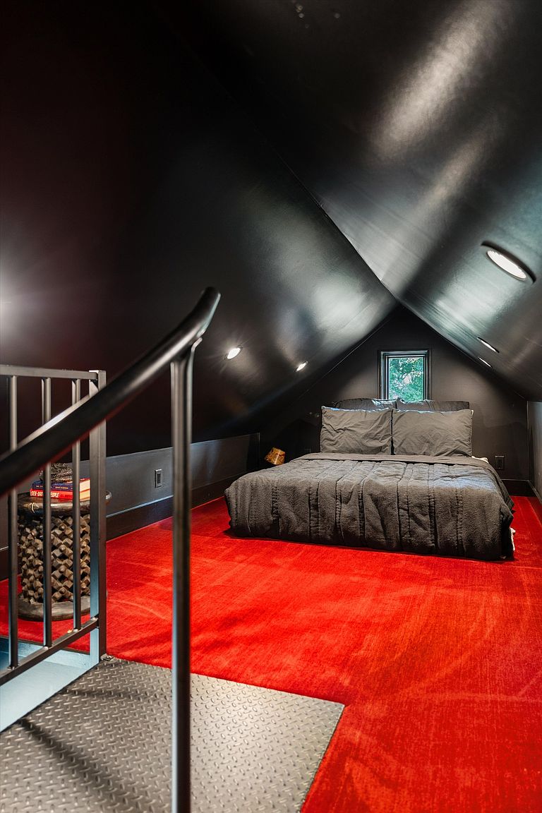 This is an interior shot of a converted attic space, likely used as a bedroom. The walls and ceiling are painted a dark color, contrasting with the bright red carpet. A bed is centered in the room, and a small window provides natural light. A metal staircase with a black railing leads up to the space, adding an industrial touch.