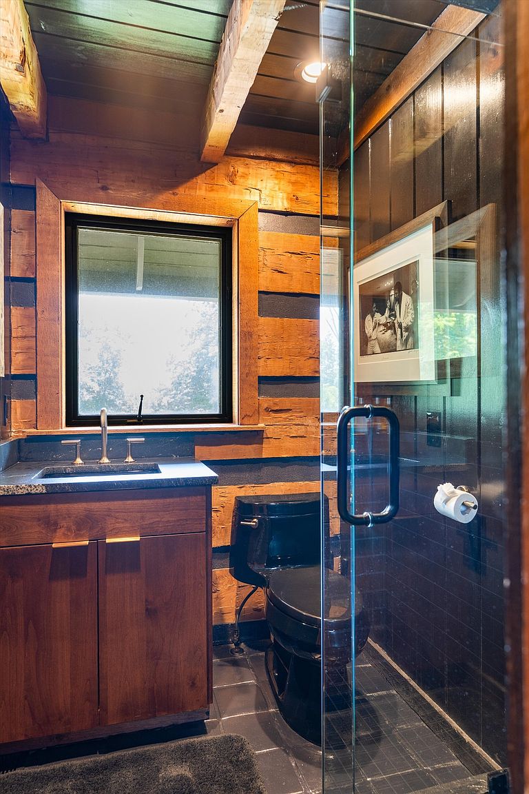 This is a rustic-style bathroom featuring wood-paneled walls, a black toilet, and a glass-enclosed shower. The vanity has a dark countertop and wooden cabinets, complemented by a black-framed window. The overall impression is a blend of modern and rustic elements, creating a unique and cozy space.