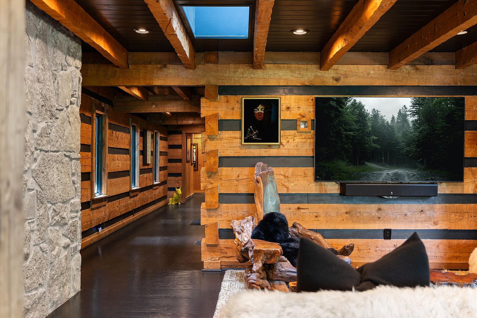 This interior shot showcases a rustic living room with exposed wooden beams and log walls, creating a warm and inviting atmosphere. A large television is mounted on the wall above a soundbar, and artwork adds a touch of sophistication. The room features a unique blend of natural elements and modern technology, perfect for a cozy retreat.