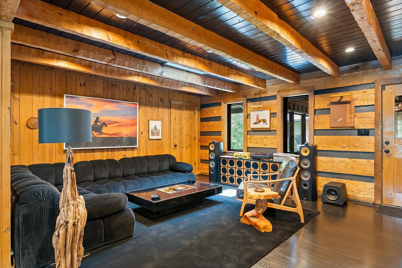 This is a cozy living room featuring wood-paneled walls and a dark wood ceiling with exposed beams. A large, comfortable sectional sofa sits on a dark rug, complemented by a unique wooden coffee table and a rustic wooden chair. The room is decorated with artwork and features a vintage-style audio system, creating a warm and inviting atmosphere.