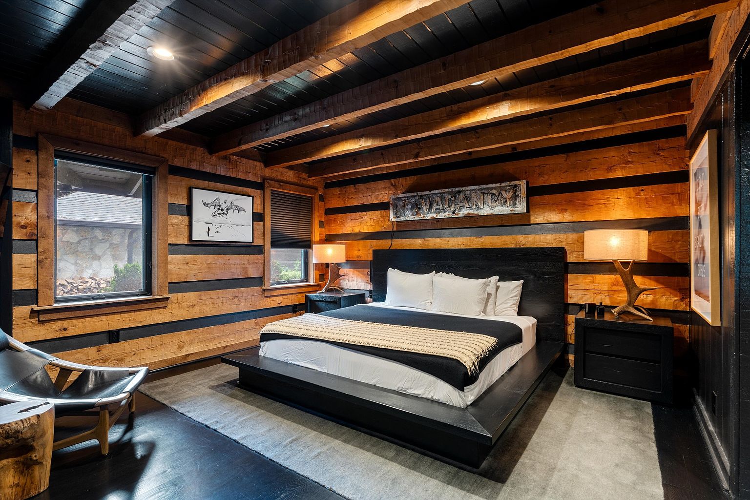 This is a primary bedroom featuring a large platform bed with white linens and a black throw blanket. The walls are wood with black accents, and the ceiling has exposed wooden beams painted black. Two nightstands with lamps flank the bed, and artwork hangs above the headboard, creating a rustic yet modern aesthetic.