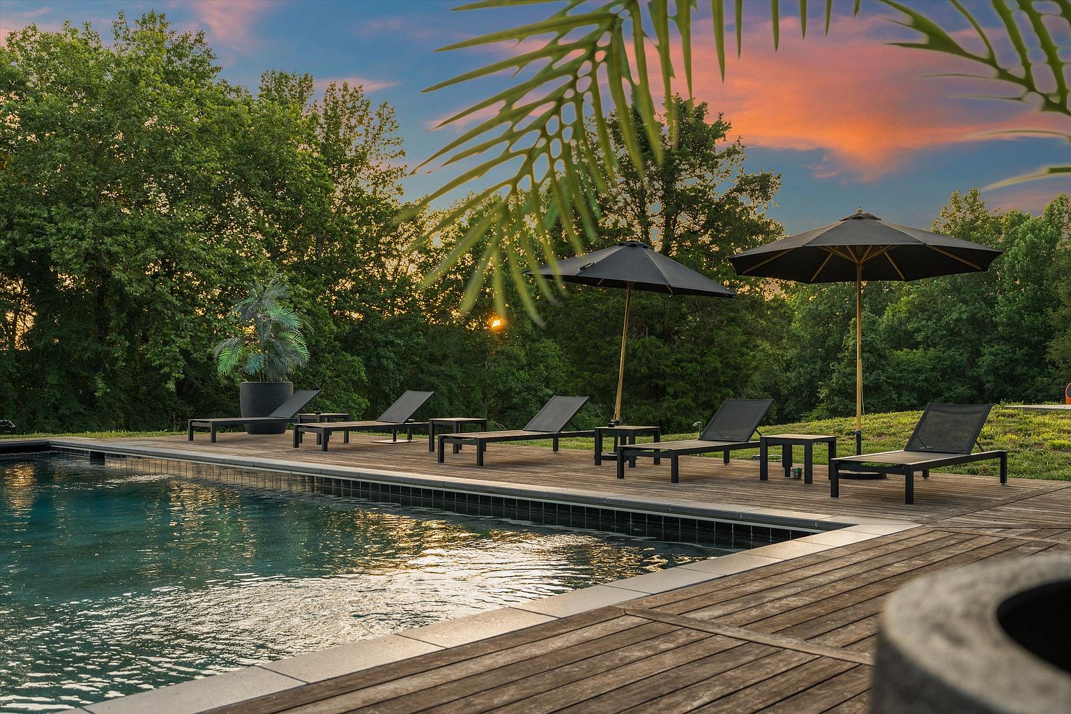 This image showcases a luxurious outdoor pool area at sunset. The pool is surrounded by a wooden deck furnished with lounge chairs and umbrellas, creating a relaxing and inviting atmosphere. Lush greenery provides privacy and enhances the serene ambiance, making it an ideal space for leisure and entertainment.