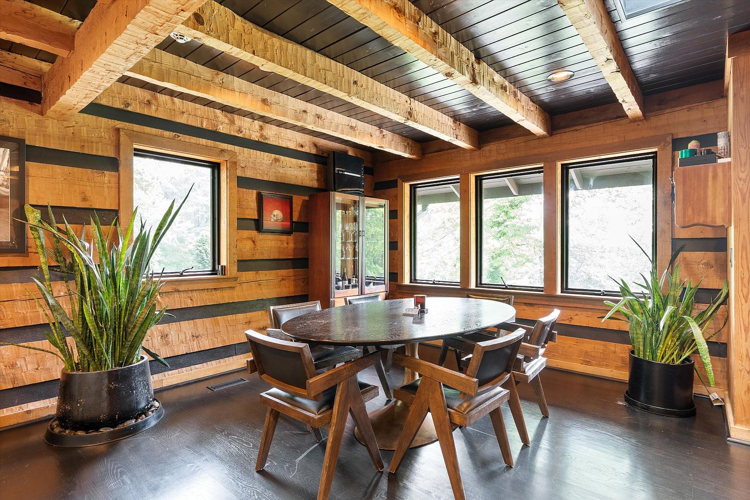 This dining room features a rustic yet modern design with wood-paneled walls and exposed beam ceilings. A dark, oval-shaped dining table is surrounded by six wooden chairs with leather seats. Large windows provide natural light, and potted plants add a touch of greenery to the space, creating a warm and inviting atmosphere.