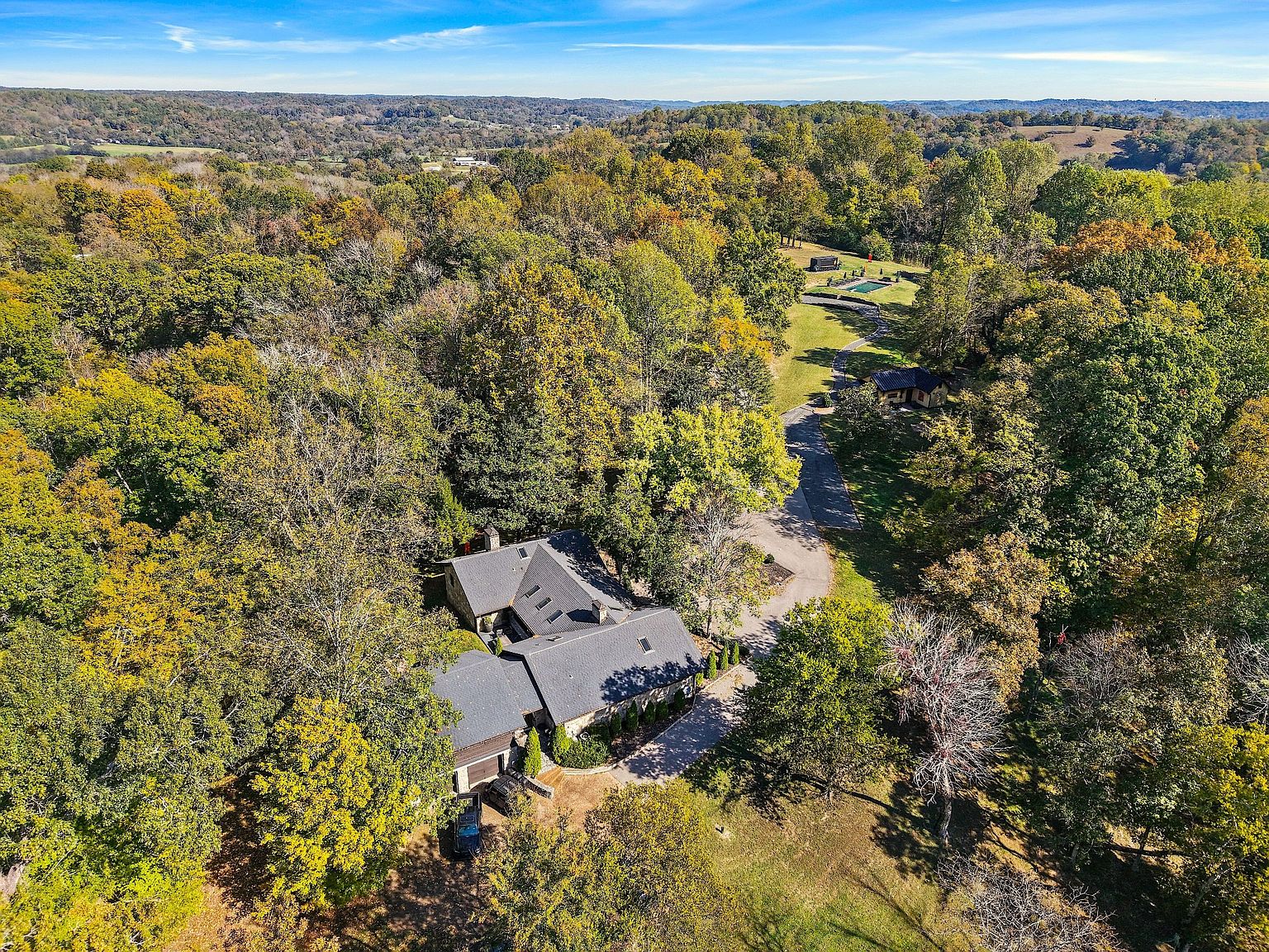 This aerial view showcases a sprawling property nestled among lush, mature trees, highlighting the home's secluded setting. The residence features a dark roof and a winding driveway, with a glimpse of a pool and additional outbuildings in the distance. The landscape is a mix of vibrant greenery and fall foliage, creating a picturesque and private estate.
