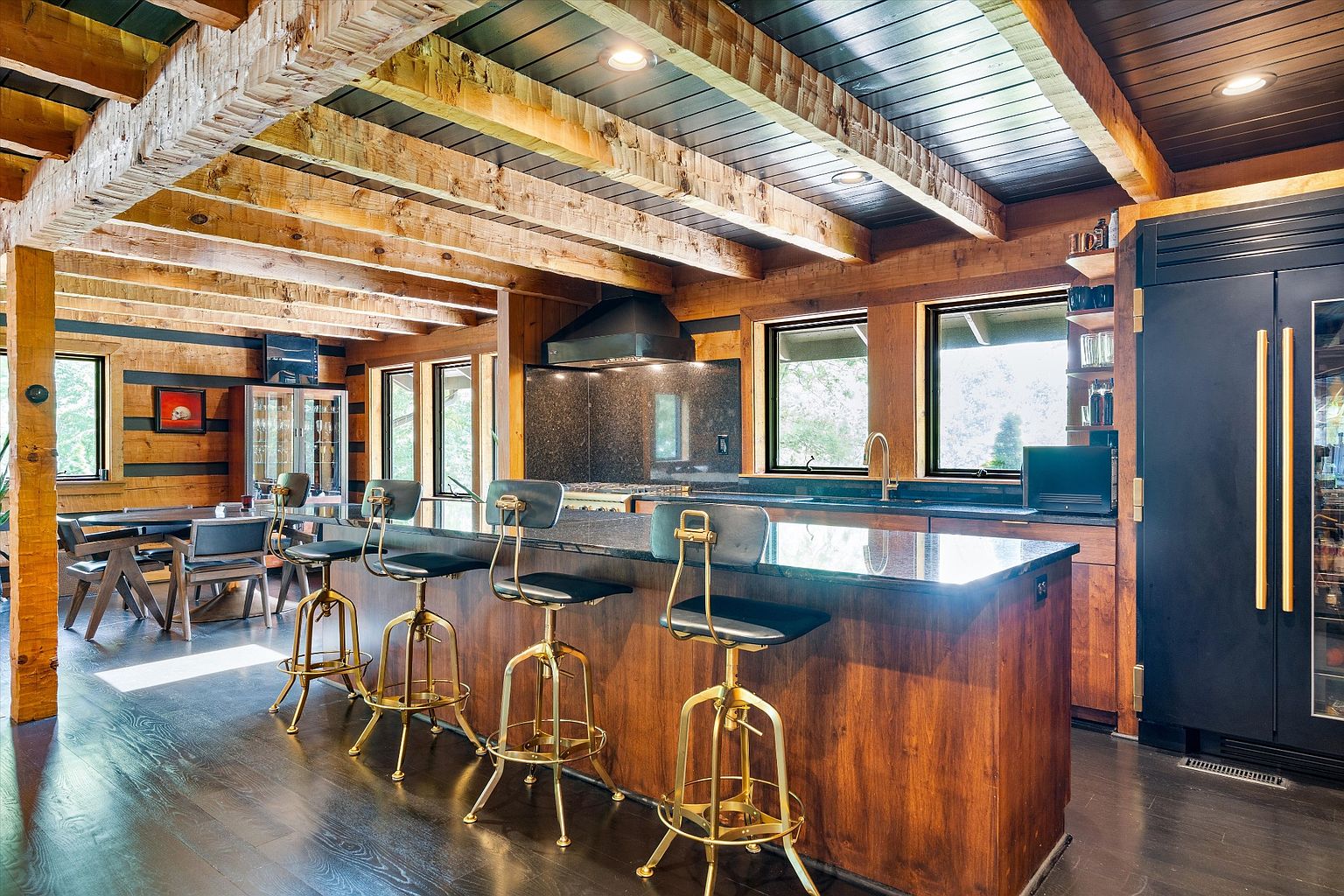 This interior shot showcases a rustic yet modern kitchen with exposed wooden beams and a dark wood floor. A kitchen island with bar stools takes center stage, complemented by sleek black appliances and dark countertops. The space is well-lit, highlighting the contrast between the natural wood and contemporary elements.