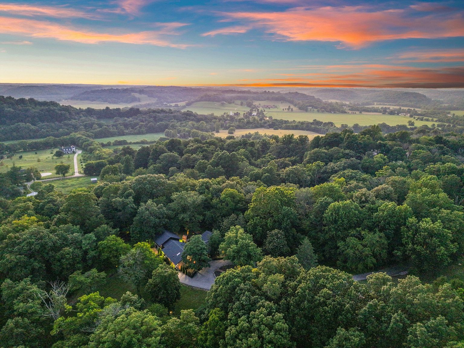 This aerial view showcases a secluded property nestled within a dense forest, offering privacy and tranquility. The home features a dark roof and is surrounded by lush greenery, with glimpses of open fields and a colorful sunset in the distance. The landscape suggests a peaceful, rural setting, ideal for those seeking a retreat.