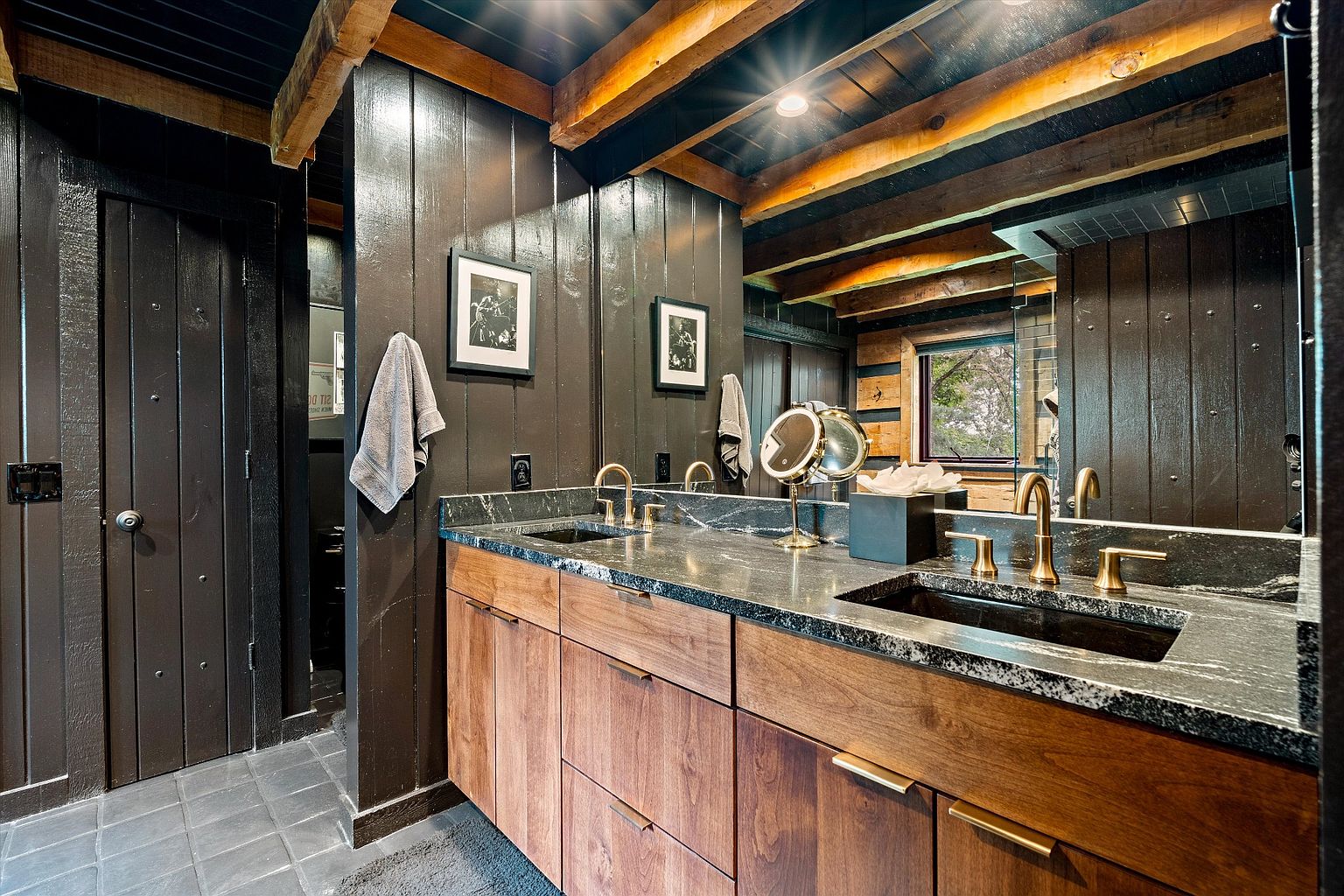 This is a primary bathroom featuring a double vanity with a dark granite countertop and wooden cabinets with gold hardware. The walls are dark wood paneling, complemented by exposed wooden beams on the ceiling. The space includes a large mirror reflecting a window and a separate shower area, creating a rustic yet luxurious feel.