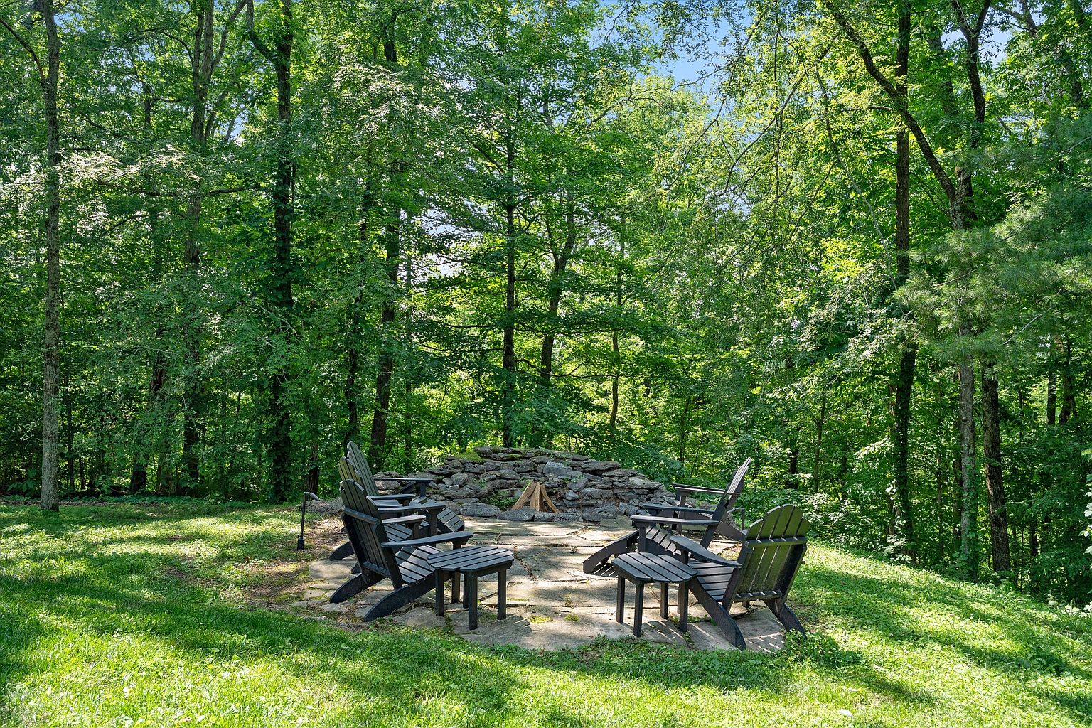 This image showcases a charming outdoor fire pit area nestled within a lush, green yard. The focal point is a circular stone fire pit surrounded by Adirondack chairs, creating an inviting space for relaxation and entertainment. The area is surrounded by mature trees, providing privacy and a serene atmosphere, making it an ideal feature for a real estate listing emphasizing outdoor living.