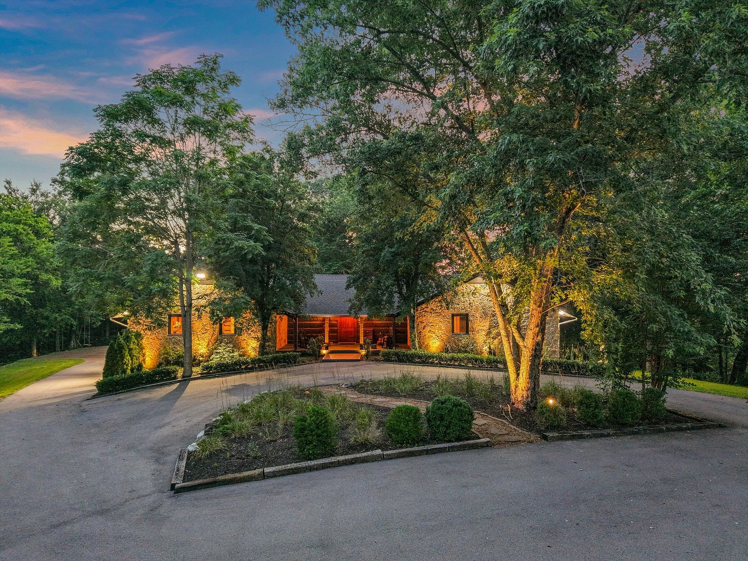 The image showcases the front exterior of a stone-clad house, partially obscured by mature trees that are illuminated by landscape lighting. A circular driveway leads to the entrance, which is framed by the trees, creating a sense of privacy and seclusion. The warm lighting highlights the architectural details and enhances the inviting atmosphere of the property.