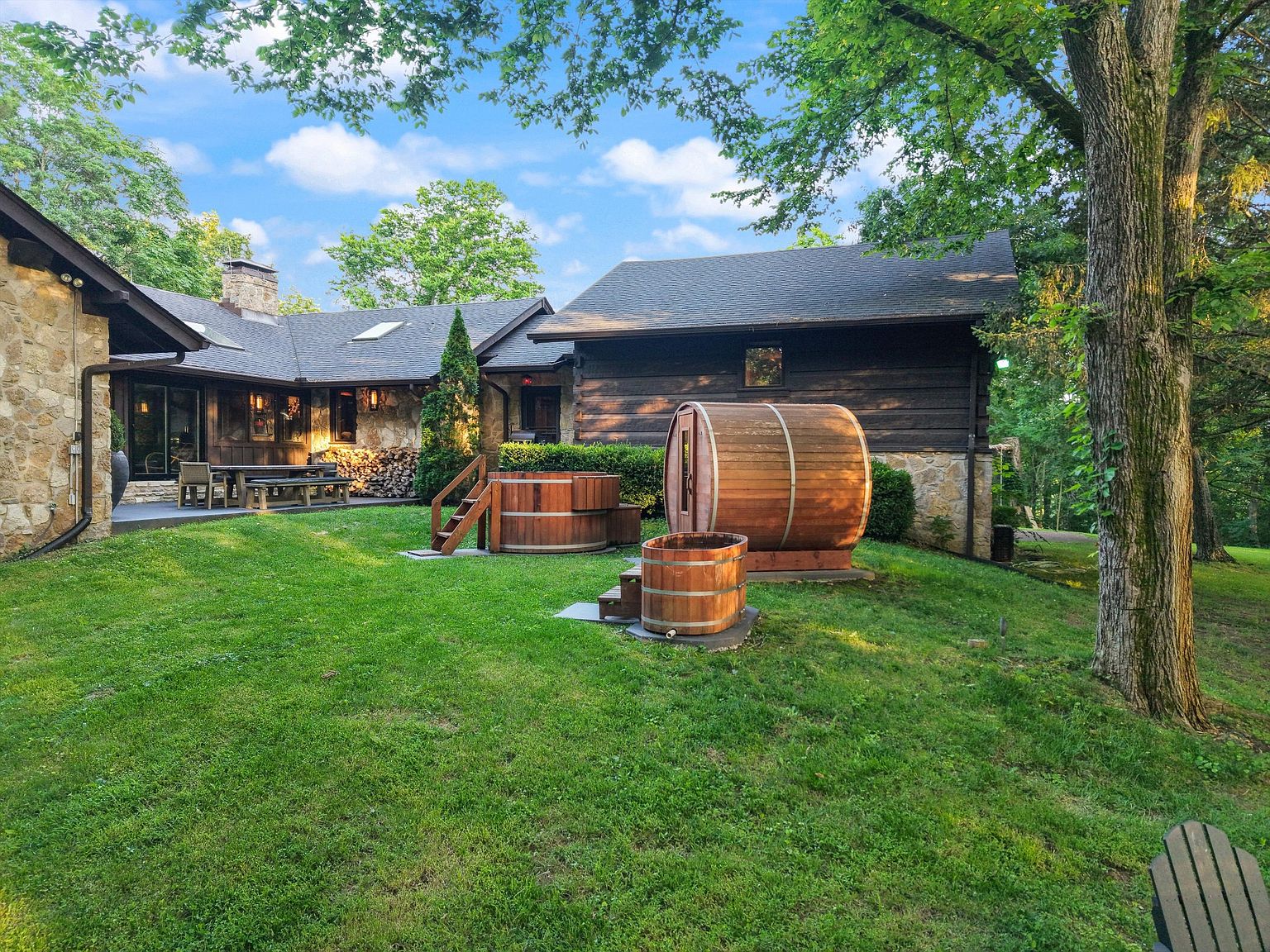 This exterior shot showcases a beautifully landscaped backyard featuring a well-maintained lawn, mature trees, and unique wooden spa features including a barrel sauna and hot tub. The stone and wood exterior of the house blends seamlessly with the natural surroundings, creating a serene and inviting atmosphere. A patio area with outdoor seating is visible, suggesting an ideal space for relaxation and entertaining.