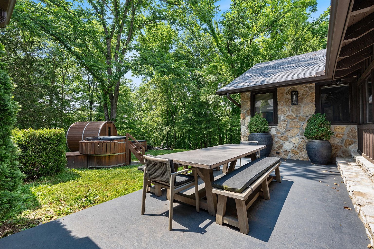 This image showcases a charming outdoor patio area, featuring a rustic wooden dining set with a long table, benches, and chairs. A wooden hot tub is visible in the background, surrounded by lush greenery and mature trees, creating a private and serene atmosphere. The patio is adjacent to a stone-clad building, adding to the overall aesthetic appeal.