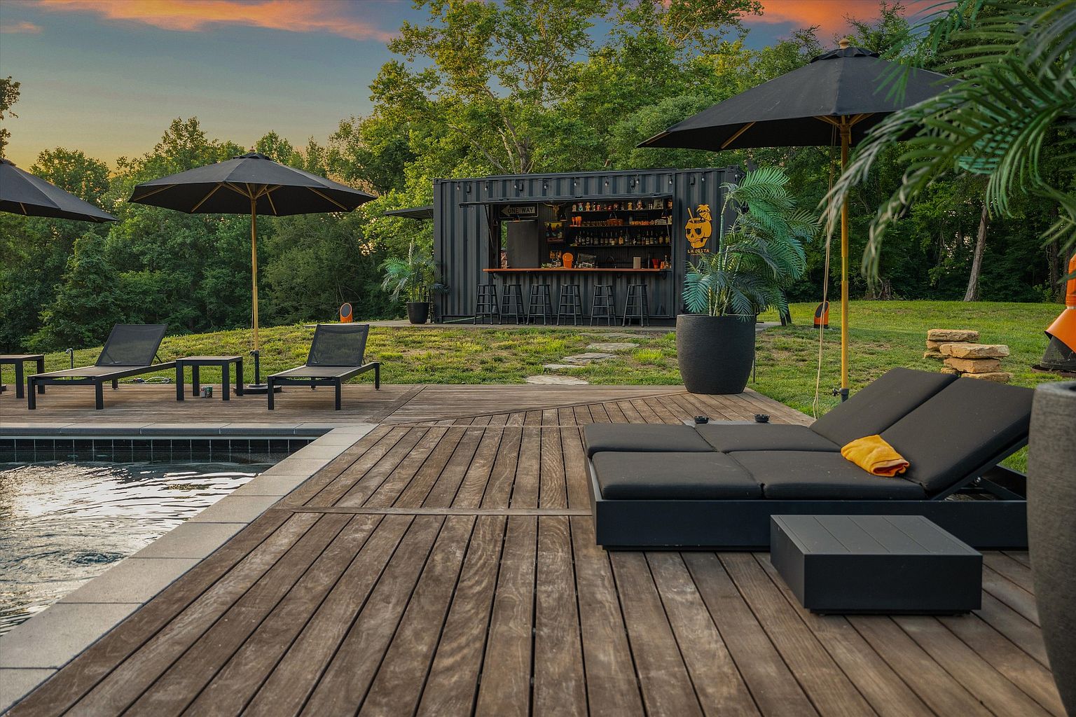 This image showcases a luxurious outdoor pool area with a modern aesthetic. The wooden deck features sleek lounge chairs and umbrellas, creating a relaxing space. A unique bar constructed from a shipping container adds a touch of industrial chic, while lush greenery provides a serene backdrop, perfect for entertaining or unwinding.