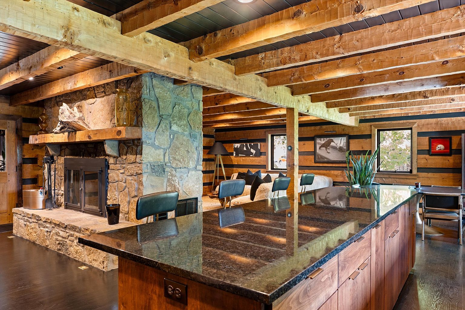 This interior shot showcases a rustic kitchen with a prominent stone fireplace and exposed wooden beams, creating a warm and inviting atmosphere. The kitchen island features a dark countertop and wooden cabinetry, complemented by modern bar stools. The open-concept design flows into a living area, enhancing the sense of spaciousness and connection within the home.