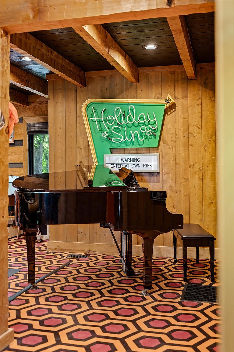 This interior shot showcases a unique living room featuring a black grand piano, a vintage-style 'Holiday Inn' sign, and distinctive geometric patterned flooring. The wood-paneled walls and ceiling beams add a rustic touch, while the piano and sign create a focal point, suggesting a space for entertainment and relaxation. The room exudes a quirky and inviting atmosphere.